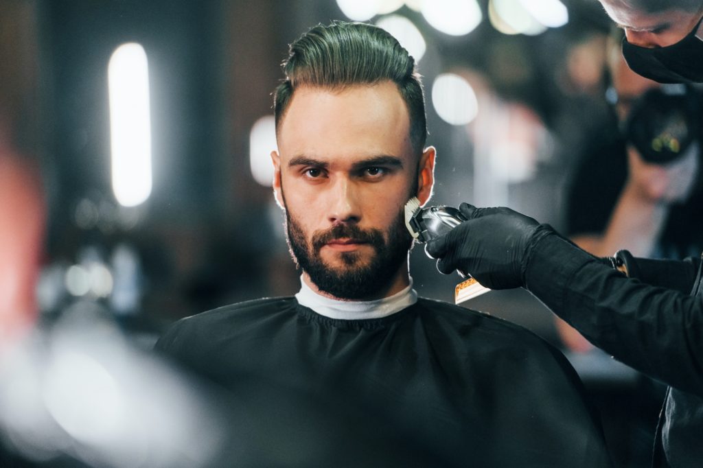 Young man with stylish hairstyle sitting and getting his beard shaved in barber shop