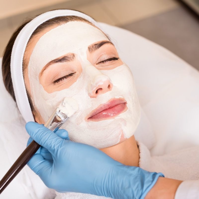 beautician applying cosmetic mask on woman face with cosmetic brush at beauty salon